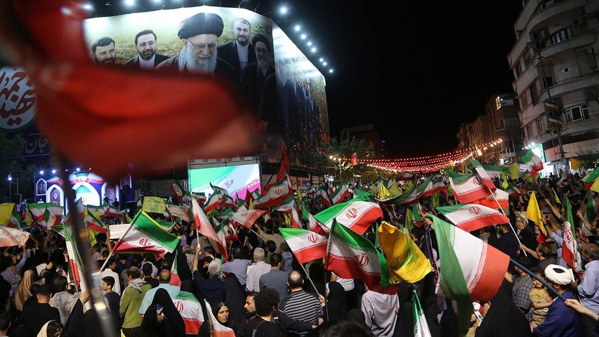Crowds wave Iranian flags at a nighttime rally in Tehran beneath a portrait of Ayatollah Khamenei.