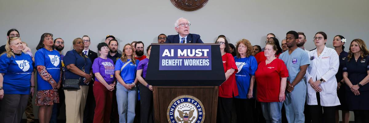 Senator Bernie Sanders speaking at a press conference podium with a sign reading 'AI MUST BENEFIT WORKERS' and union workers standing behind him.
