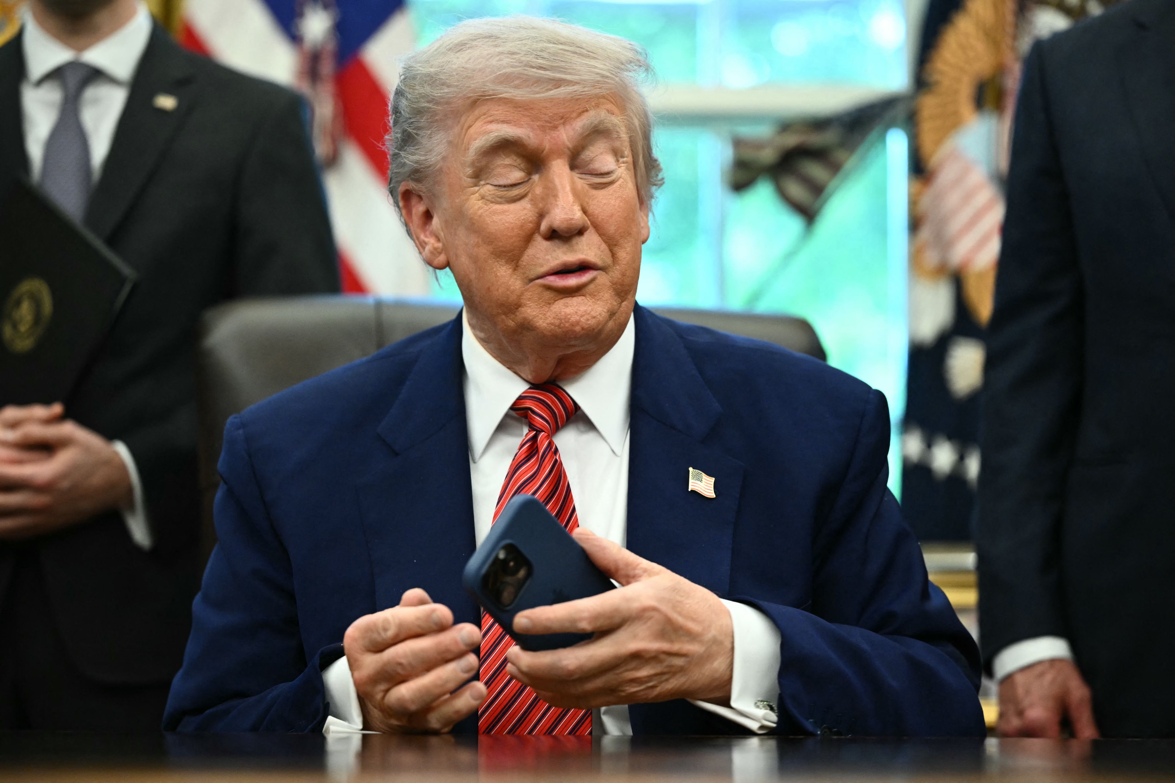 President Donald Trump speaking from behind the Resolute Desk in the Oval Office, holding a phone.