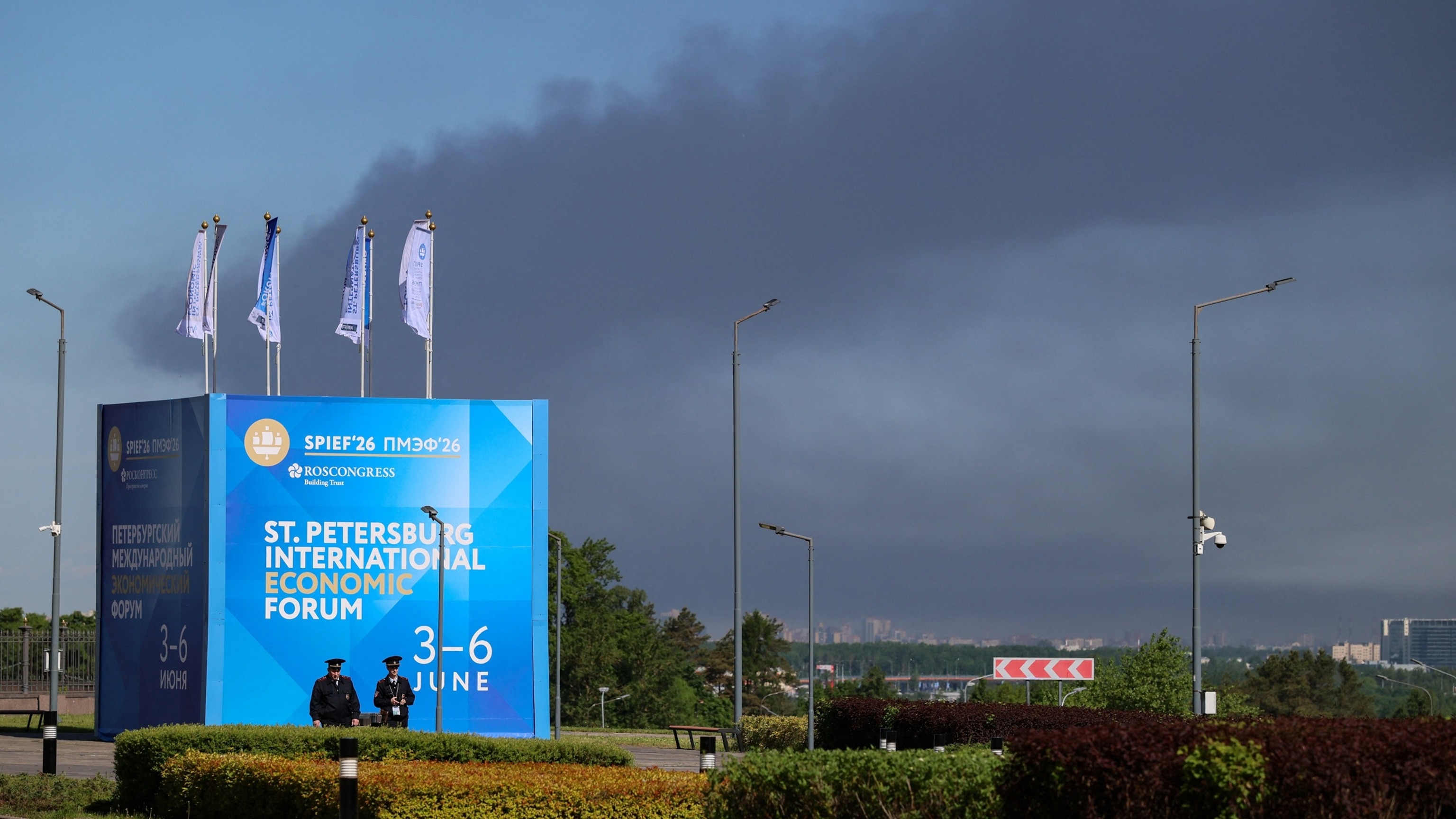 Police officers walk past a SPIEF 2026 banner as black smoke from the burning oil terminal drifts across the sky behind them.
